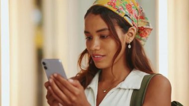 Uzun kahverengi saçlı, beyaz bluzlu ve sırtında sırt çantası olan sarı bandanalı hoş, bronz tenli kadın modern bina arka planında cep telefonu kullanıyor..