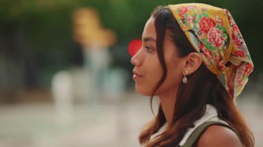 Close-up profile of cute tanned woman with long brown hair wearing white top and yellow bandana walking on crosswalk on passing cars blurred background