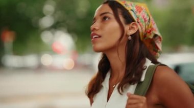 Close-up profile of cute tanned woman with long brown hair wearing white top and yellow bandana walking on crosswalk on passing cars blurred background
