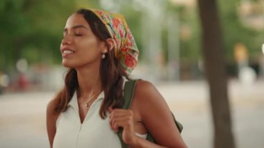 Cute tanned woman with long brown hair wearing white top and yellow bandana walking on crosswalk on passing cars blurred background