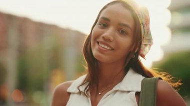 Clouse-up, cute tanned woman with long brown hair wearing white top and yellow bandana posing on camera. Smiling girl looking at the camera. Backlight