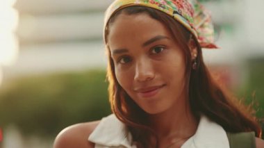Clouse-up, cute tanned woman with long brown hair wearing white top and yellow bandana posing on camera. Smiling girl looking at the camera. Backlight