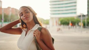 Clouse-up, cute tanned woman with long brown hair wearing white top and yellow bandana posing on camera. Smiling girl looking at the camera. Girl smiles and puts her hand to her hair. Backlight