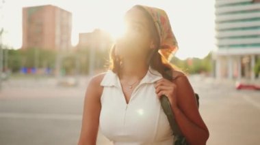 Cute tanned woman with long brown hair wearing white top and yellow bandana walks down the street modern building background. Backlight. Camera moving around