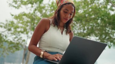 Cute tanned woman with long brown hair in white top and yellow bandana using laptop while sitting on bench. Pretty girl student preparing for classes in outside