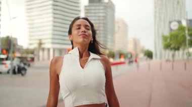 Gülümseyen, bronz tenli, uzun kahverengi saçlı, beyaz bluzlu, esintiden keyif alan hoş bir kadının yakın çekimi. Kamera ileri gidiyor.