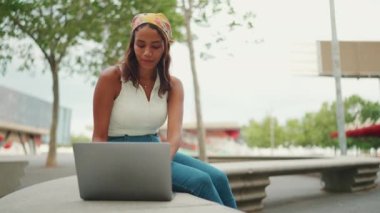 Cute tanned woman with long brown hair in white top and yellow bandana using laptop while sitting on bench. Pretty girl student preparing for classes in outside