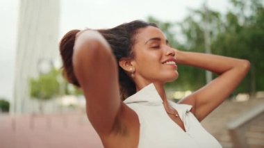 Gülümseyen, bronz tenli, uzun kahverengi saçlı, beyaz bluzlu güzel bir kadın ellerini kaldırıyor ve saçlarına dokunuyor. Kız esintiden hoşlanıyor. Kamera ileri gidiyor.