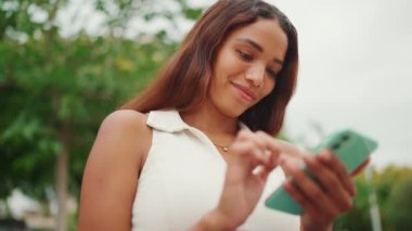 Uzun siyah saçlı, beyaz bluzlu güzel kız bankta oturuyor ve cep telefonu kullanıyor. Sosyal ağlarda cep telefonuyla yazan gülümseyen genç bir kadın.