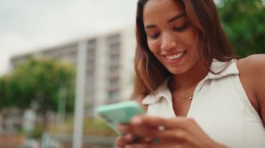 Clouse-up, beautiful girl with long dark hair wearing white top sits on bench and uses mobile phone. Young smiling woman writing in social networks on cellphone. Sideways movement