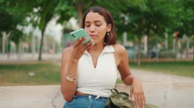 Beautiful girl with long dark hair wearing white top sits on bench and uses mobile phone. Young woman sends voice message