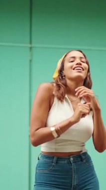 Uzun kahverengi saçlı, beyaz bluzlu ve sarı bandanalı hoş bir kadın kablosuz kulaklıkla müzik dinliyor ve mavi duvar arkasında dans ediyor.