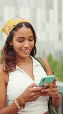 Beyaz bluzlu ve sarı bandanalı hoş bir kadın sırtında sırt çantası ve elinde cep telefonuyla köprüde yürüyor ve modern şehir hayatına bakıyor.