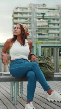 Beyaz bluzlu, uzun kahverengi saçlı, sevimli bir kadın ve şehir manzarası arka planında bankta oturan sarı bandana. Kamera ileri gidiyor.