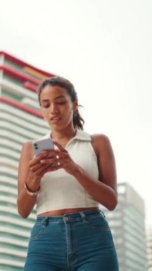 VERTICAL VIDEO: Cute tanned woman with ponytail wearing white top looking up at street signs and map trying to find her way using cellphone. Girl using map application outdoors