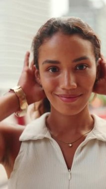 Yakından bronzlaşmış, koyu renk saçlı ve kahverengi gözlü güzel bir kadın kameraya gülümseyerek bakıyor, ellerini kaldırıyor, saçlarını düzleştiriyor.