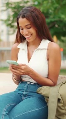 Uzun siyah saçlı, beyaz bluzlu güzel bir kız bankta oturuyor ve cep telefonu kullanıyor. Sosyal ağlarda cep telefonuyla yazan gülümseyen genç bir kadın.