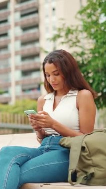 Uzun siyah saçlı, beyaz bluzlu güzel bir kız bankta oturuyor ve cep telefonu kullanıyor. Sosyal ağlarda cep telefonuyla yazan gülümseyen genç bir kadın.