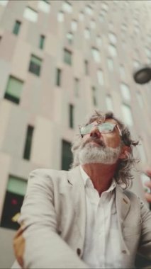 VERTICAL VIDEO: Clouse up, mature businessman with beard in eyeglasses wearing gray pondering jacket looks around