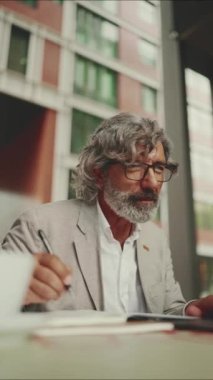Gözlüklü, sakallı, dizüstü bilgisayarlı ve not alan, açık kafede oturan olgun bir iş adamı. Mobil ofis çalışanı.
