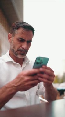 Beyaz gömlek giyen, sakallı, olgun bir iş adamı cep telefonu kullanıyor. Kafe masasında oturan başarılı adam, akıllı telefondan yazı yazıyor.