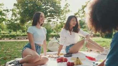 Happy smiling young multinational people at picnic on summer day outdoors. Group friends talking while spending the weekend together, relaxing in the park at picnic