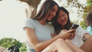 Young multi ethnic smiling women are watching videos on their phone. Girls laugh at picnic on summer day outdoors