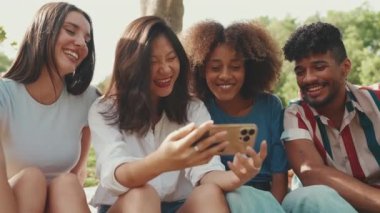 Yazın açık havada cep telefonuyla video izleyen mutlu, etnik çeşitliliğe sahip genç kadınların yakın çekimi. Bir grup arkadaş akıllı telefon kullanır, video izler, sosyal ağlar