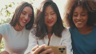 Yazın açık havada cep telefonuyla video izleyen mutlu, etnik çeşitliliğe sahip genç kadınların yakın çekimi. Bir grup arkadaş akıllı telefon kullanır, video izler, sosyal ağlar