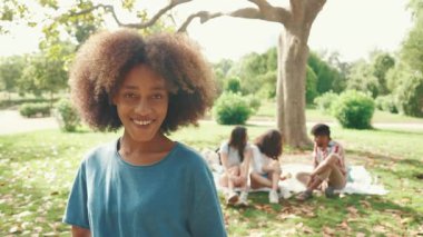 Kıvırcık saçlı, mavi tişörtlü, parkta kameraya poz veren genç bir kadının yakın çekimi. Yaz günü açık havada piknik. Arkadaşları arka planda bulanık oturuyor.