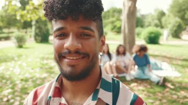 Kıvırcık saçlı, çizgili gömlek giyen ve parkta kameraya poz veren genç bir adamın portresi. Yaz günü açık havada piknik. Arkadaşları arka planda bulanık oturuyor.