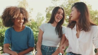 Yaz günü açık havada parkta bankta oturan mutlu çoklu etnik kökenli genç kadınlar konuşuyor. Şehir parkında bir grup kız neşeyle konuşuyor ve gülüyor.
