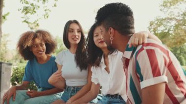 Yaz günü açık havada parkta bankta oturan mutlu çoklu etnik gruptan gençler konuşuyor. Şehir parkında bir grup arkadaş neşeyle konuşuyor ve gülüyor.