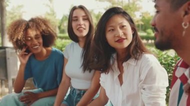 Yaz günü açık havada parkta bankta oturan mutlu çoklu etnik gruptan gençler konuşuyor. Şehir parkında bir grup arkadaş neşeyle konuşuyor ve gülüyor.