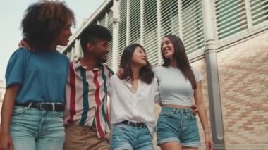 Happy multiethnic young people walk embracing on summer day outdoors. Group of friends are talking and laughing merrily while walking along on the street