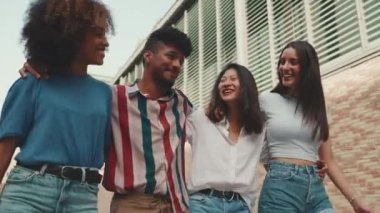 Happy multiethnic young people walk embracing on summer day outdoors. Group of friends are talking and laughing merrily while walking along on the street