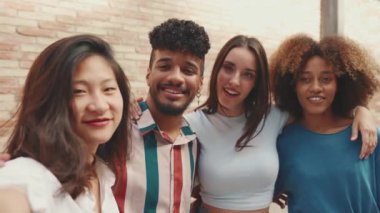Mutlu, çok ırklı genç insanlar dışarıda yaz günü fotoğraf çektiriyorlar. Bir grup arkadaş cep telefonu kamerasına sıkıca gülümserken tuğla duvar arkasında duruyorlar.