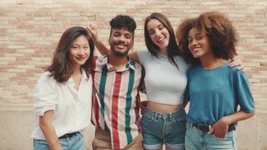 Happy, lovely multiethnic young people posing for the camera on summer day outdoors. Group of friends hugging tightly smiling at camera while standing on brick wall background