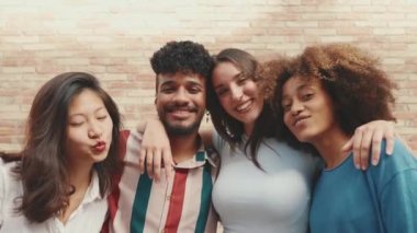 Happy, lovely multiethnic young people posing for the camera taking selfie on summer day outdoors. Group of friends hugging tightly smiling at cellphone camera while standing on brick wall background