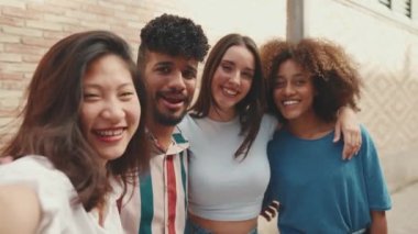 Happy, lovely multiethnic young people posing for the camera taking selfie on summer day outdoors. Group of friends hugging tightly smiling at cellphone camera while standing on brick wall background