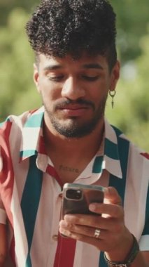 Kıvırcık saçlı, çizgili tişörtlü, gülümseyen, yaz günü parkta piknik yapan arkadaşlarıyla konuşan, cep telefonu kullanan genç bir erkek.