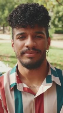 Kıvırcık saçlı, çizgili gömlekli, parkta kameraya poz veren gülümseyen genç adamın yakın çekimi. Açık havada piknik.