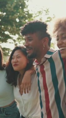 VERTICAL VIDEO: Happy multiethnic young people walk embracing on summer day outdoors. Group of friends are talking and laughing merrily while walking along path in city park