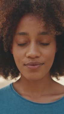 Kıvırcık saçlı, mavi tişörtlü ve parkta kameraya poz veren genç bir kadının yakın çekimi. Kız gözlerini açar ve kameraya gülümser.