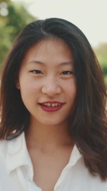 Parkta kameraya poz veren uzun kahverengi saçlı, uzun boylu, gülümseyen Asyalı bir kadın. Kız saçına dokunur ve kameraya gülümser.