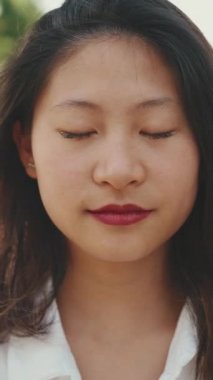 Parkta kameraya poz veren uzun kahverengi saçlı, uzun boylu, gülümseyen Asyalı bir kadın. Kız gözlerini açar ve kameraya gülümser.