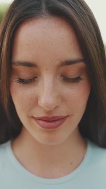 Parkta kameraya poz veren uzun kahverengi saçlı, beyaz ekin bluzlu, gülümseyen genç bir kadının yakın çekimi. Kız gözlerini açar ve kameraya gülümser.