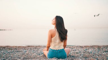 Kumsalda deniz manzaralı, uzun saçlı güzel bir kadın oturuyor. Üstü başı ve kot şortu olan güzel bir kız.. 