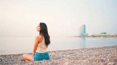 Kumsalda deniz manzaralı, uzun saçlı güzel bir kadın oturuyor. Üstü başı ve kot şortu olan güzel bir kız.. 