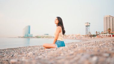 Kumsalda deniz manzaralı, uzun saçlı güzel bir kadın oturuyor. Üstü başı ve kot şortu olan güzel bir kız.. 